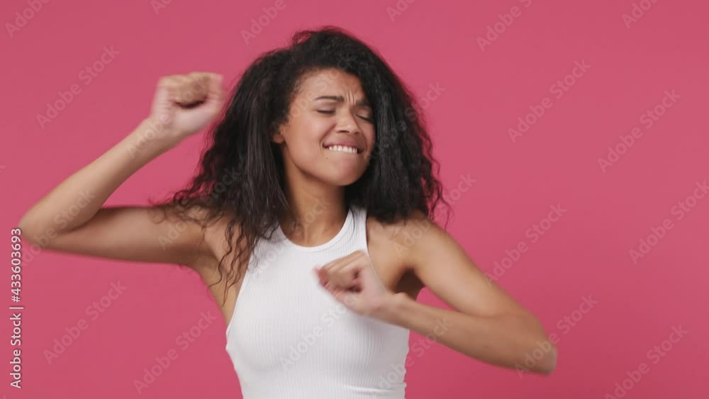 Young african woman curly hair wears white tank top shirt dance clenching fists waving rising gesticulating hands fool around have fun enjoy celebrate play isolated on dark pink color wall background Young african woman curly hair wears white tank top shirt dance clenching fists waving rising gesticulating hands fool around have fun enjoy celebrate play isolated on dark pink color wall background