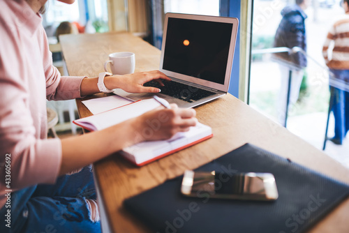 Crop woman taking notes and browsing laptop