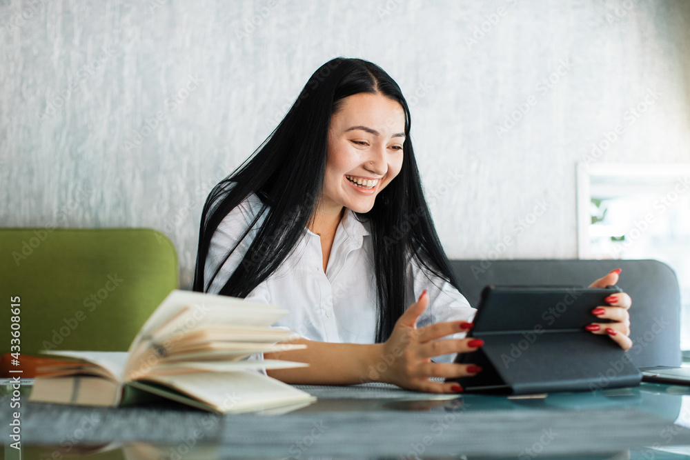 Happy joyful likable Asian woman uses digital tablet, while sitting at the table in coffee shop. Young businesswoman having video call, smiling cheerfully to webcamera Happy joyful likable Asian woman uses digital tablet, while sitting at the table in coffee shop. Young businesswoman having video call, smiling cheerfully to webcamera