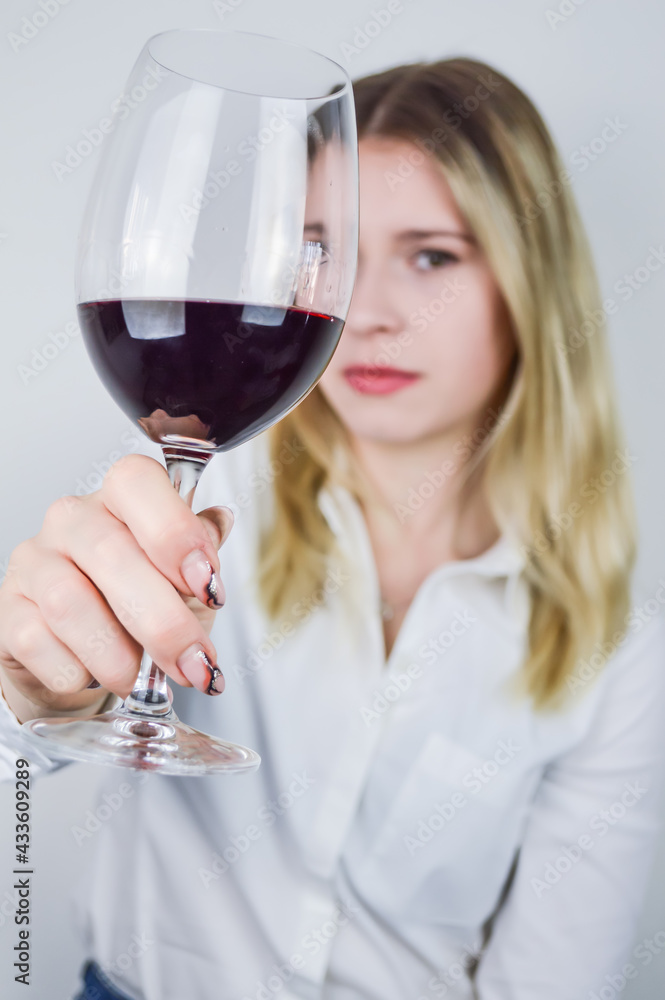 Portrait of a beautiful young blonde woman who raises a glass of red wine to appreciate its color and nuances at a wine tasting - Focus on the glass of wine Portrait of a beautiful young blonde woman who raises a glass of red wine to appreciate its color and nuances at a wine tasting - Focus on the glass of wine