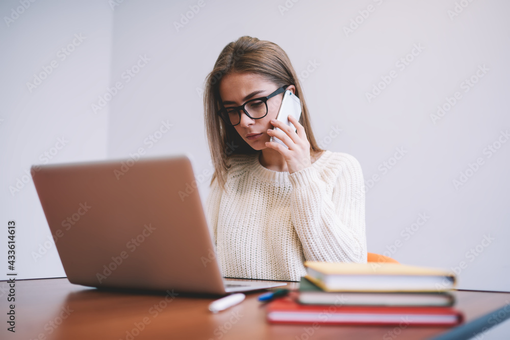 Focused young female employee speaking via smartphone while working on laptop Focused young female employee speaking via smartphone while working on laptop