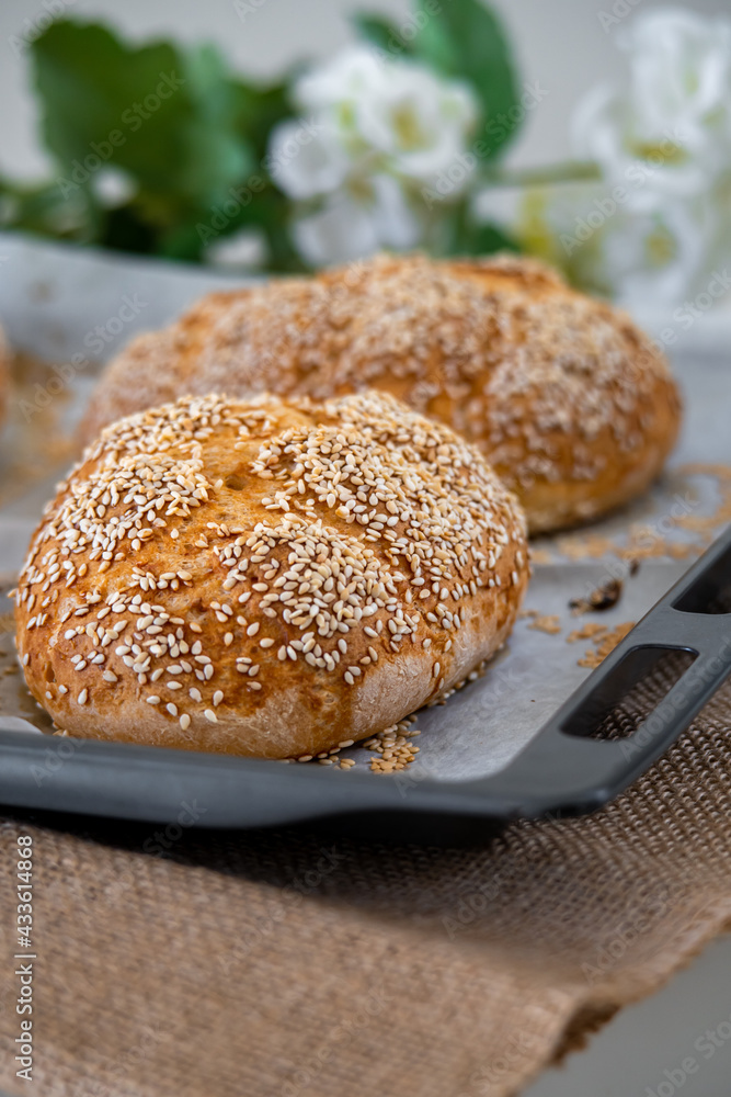 Fresh challah bread rolls hot from the oven on baking tray. Gluten free bread buns or mini loaves with sesame seeds and golden color ready to eat. Fluffy shaped homemade bread, flowers in background. Fresh challah bread rolls hot from the oven on baking tray. Gluten free bread buns or mini loaves with sesame seeds and golden color ready to eat. Fluffy shaped homemade bread, flowers in background.