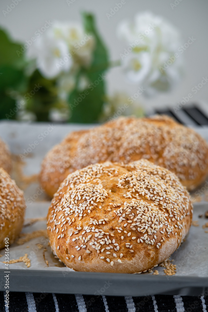 Fresh challah bread rolls hot from the oven on baking tray. Gluten free bread buns or mini loaves with sesame seeds and golden color ready to eat. Fluffy shaped homemade bread, flowers in background.