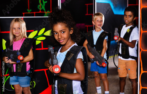 Portrait of african preteen girl standing with laser gun on the dark lasertag arena on background with her team