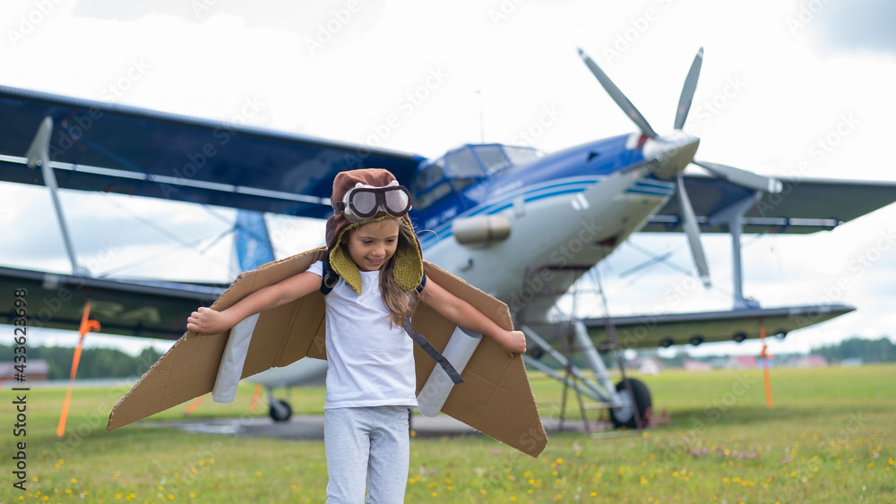 A little girl in a pilot's costume with cardboard wings runs on the lawn against the backdrop of the plane. A child in a hat and glasses dreams of flying on an airplane. A little girl in a pilot's costume with cardboard wings runs on the lawn against the backdrop of the plane. A child in a hat and glasses dreams of flying on an airplane.