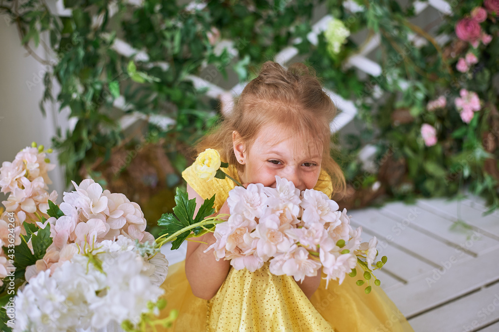 Girl, 7, sits against a green wall with flowers in a yellow dress and sniffs a white flower Girl, 7, sits against a green wall with flowers in a yellow dress and sniffs a white flower