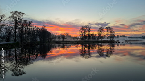 orange sunset over the lake covered with fog. The lake water is calm and forms a mirror. Perfect reflection in the water in which the orange sunset sky and tree silhouettes on the lake shore.