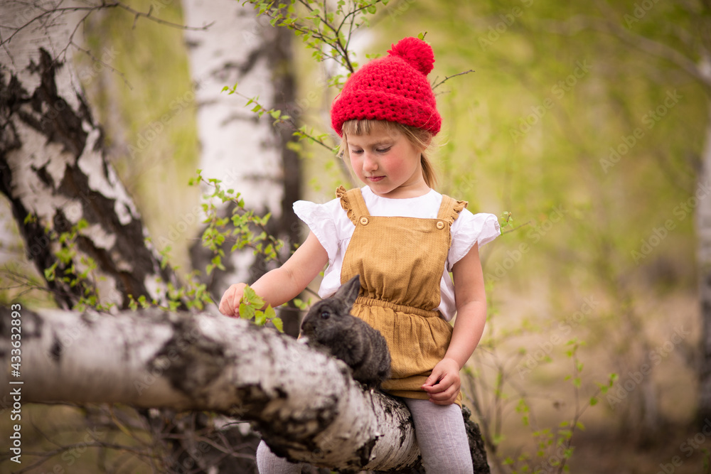 Little girl in a red hat in a spring forest plays with a rabbit in nature Little girl in a red hat in a spring forest plays with a rabbit in nature