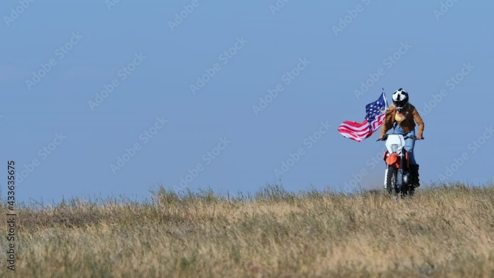 Woman with fixed American flag rides sports bike on dry grass field under clear blue sky on spring day super slow motion low angle shot Woman with fixed American flag rides sports bike on dry grass field under clear blue sky on spring day super slow motion low angle shot