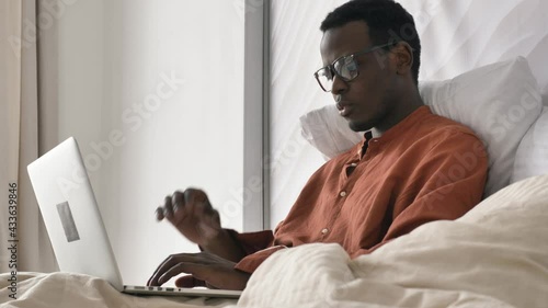 Concentrated African-American man with glasses in pajama works on laptop recovering after covid on comfortable bed in room closeup
