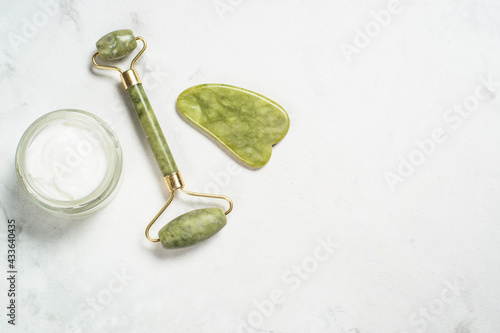 Fototapeta Naklejka Na Ścianę i Meble -  Jade roller and gua sha stone massager with face cream at white marble background. Trendy cosmetic products. Anti-aging therapy. Top view.
