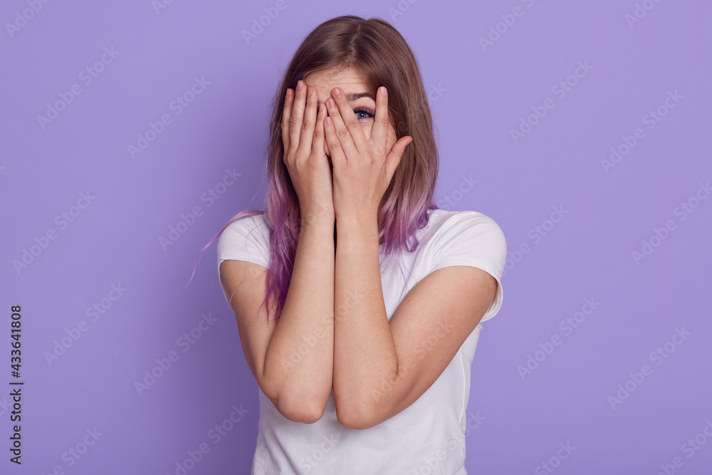 Attractive young woman wearing white casual t shirt covering her face with palms and looking through fingers, spying for somebody, isolated over purple background. Attractive young woman wearing white casual t shirt covering her face with palms and looking through fingers, spying for somebody, isolated over purple background.