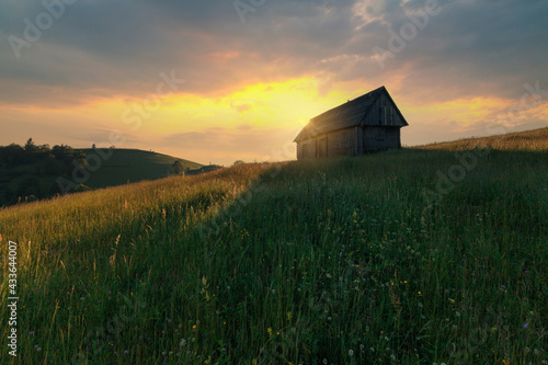 Dramatic view of high grass mountain hill under sunset clouds with the backlighted old wooden rustic house. Summer landscape.