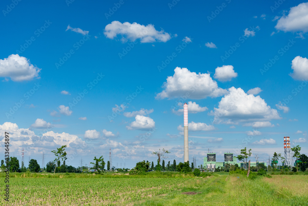 Thermoelectric power plant on the horizon, in a rural environment, with green cultivated field used in agriculture. Blue sky with white clouds on the background, Italy.