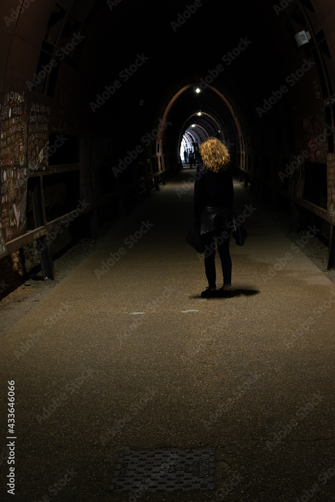 Womam in low key on grainy ground, with blonde curly hair is standing in the middle of a pedestrian dark tunnel, in a spot light. Albisola, Liguria, Italy.