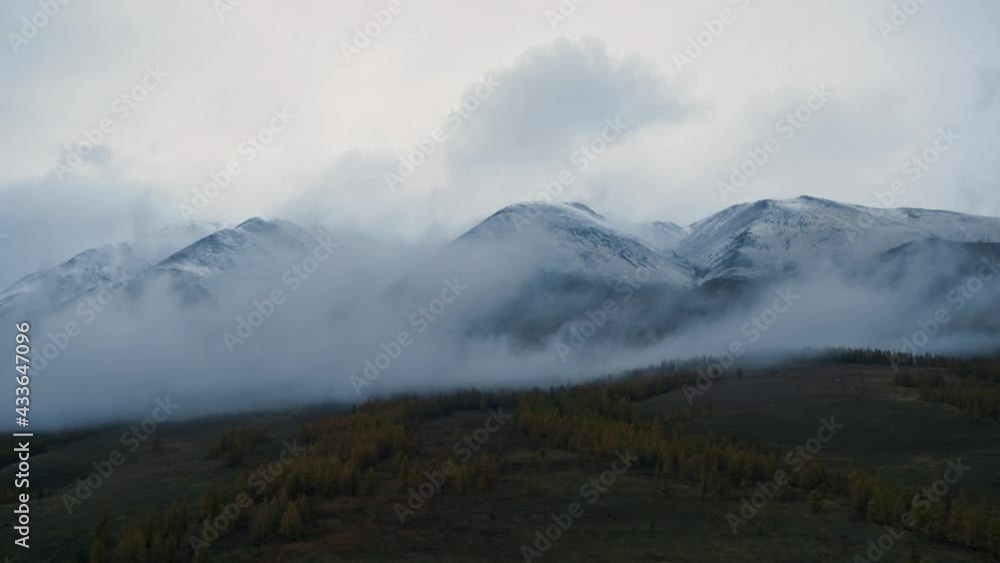 Fog over snowy mountain peaks. Drone view of magnificent landscape of Altay mountains with fog flowing over snow covered peaks and forested hills in twilight. Clouds touch mountains Fog over snowy mountain peaks. Drone view of magnificent landscape of Altay mountains with fog flowing over snow covered peaks and forested hills in twilight. Clouds touch mountains