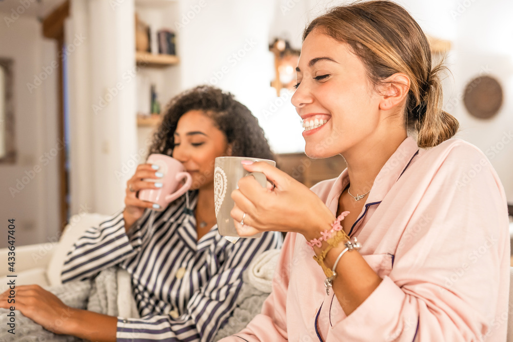 Couple of cohabiting girls in pajamas relaxing under blanket on cozy sofa drink a cup of tea while having fun talking and joking with each other. Real life moments of diverse multiracial gay couple Couple of cohabiting girls in pajamas relaxing under blanket on cozy sofa drink a cup of tea while having fun talking and joking with each other. Real life moments of diverse multiracial gay couple