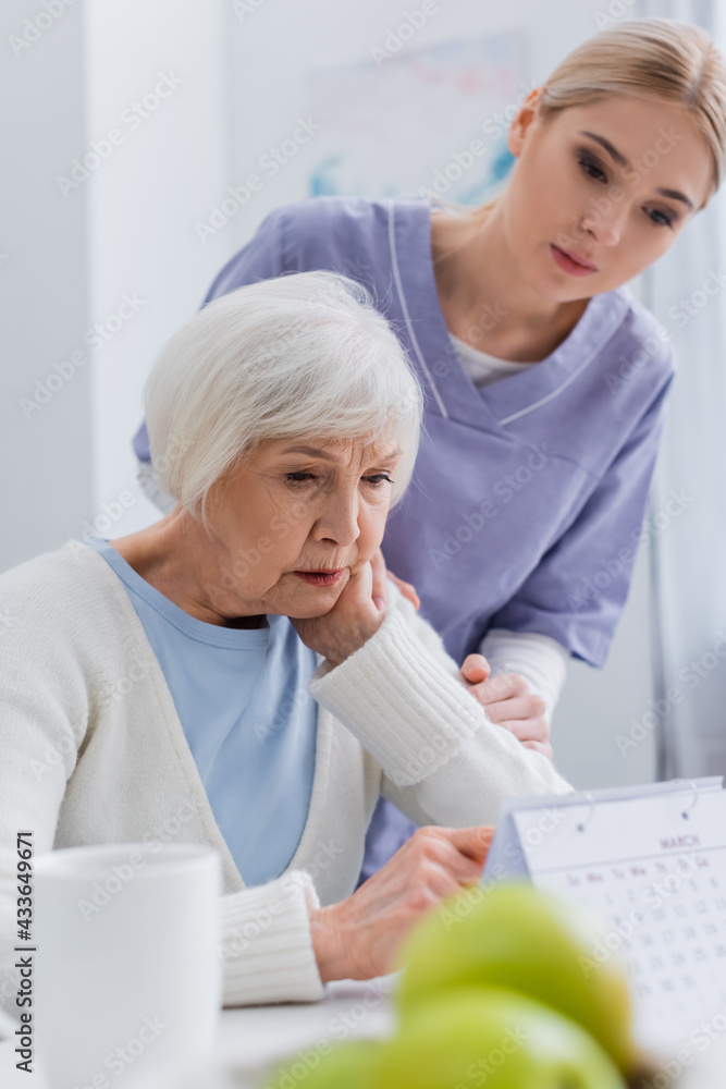 Fototapeta premium young social worker near aged woman looking at calendar on blurred foreground
