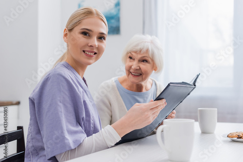 happy social worker looking at camera while holding photo album near aged woman © LIGHTFIELD STUDIOS