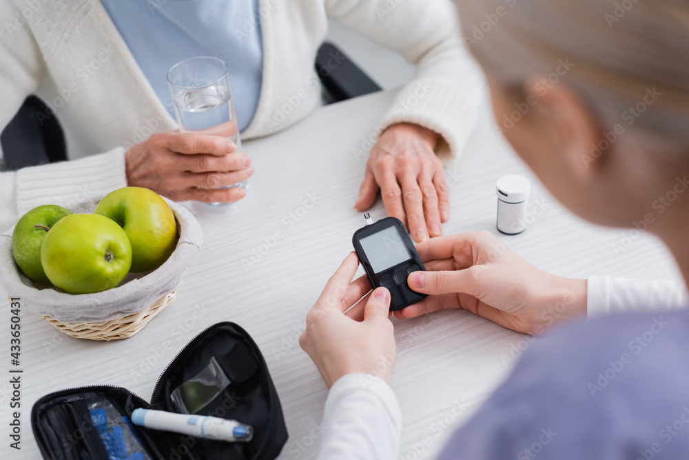 cropped view of social worker with glucometer near elderly diabetic woman with glass of water, blurred foreground
