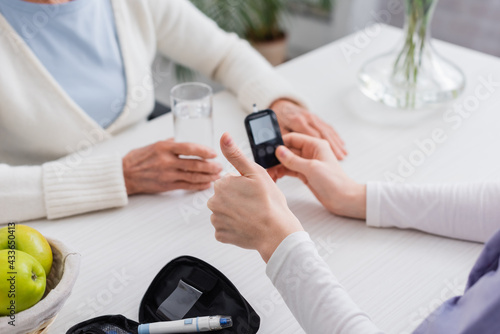 cropped view of social worker showing thumb up near glucometer and senior diabetic woman