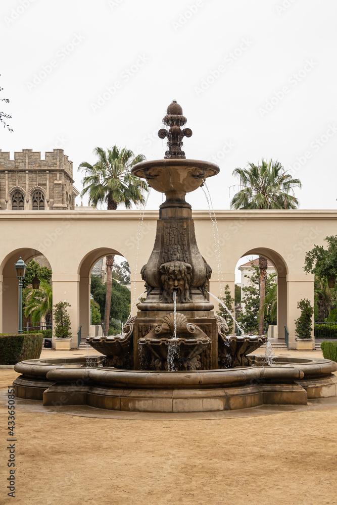 Obraz premium Pasadena, CA, USA - May 11, 2021: Bronze fountain on sandy courtyard and green foliage garden of historic City Hall under silver sky. Beige stone constructions in back.