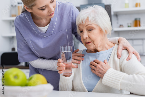 social worker hugging shoulders of aged woman holding glass of water and touching chest, blurred foreground