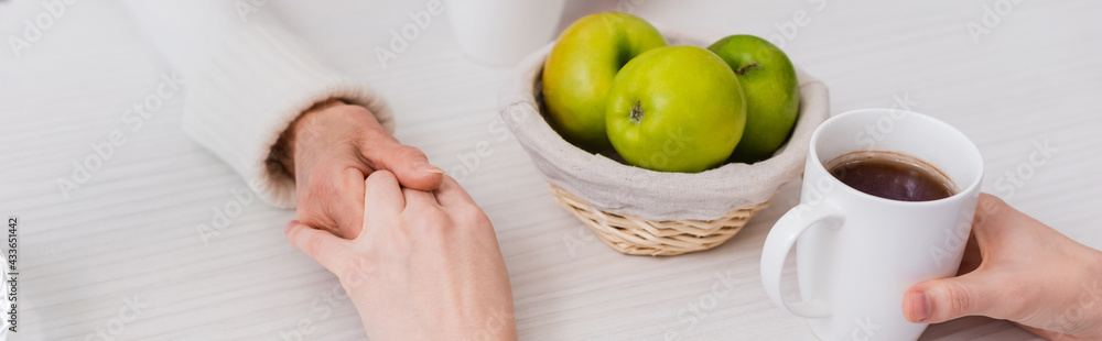 partial view of social worker and elderly woman holding hands near ripe apples and cup of tea, banner partial view of social worker and elderly woman holding hands near ripe apples and cup of tea, banner