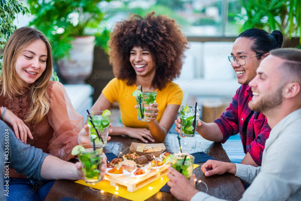 Multiracial group of friends enjoying mojito in a bar restaurant at sunset in summer making a toast. Young people holding cocktail with food cheering. Friendship, youth, social and lifestyle concept - obrazy, fototapety, plakaty 