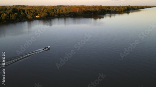 Aerial shot of cruising speed boat on calm amazon river approaching camera during epic sunset at background.