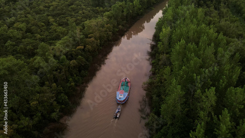 Shot of ship carrying small boat on amazon river surrounded by green rainforest trees during sunset.