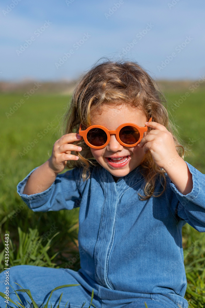 Cute happy little girl in trendy clothes and sunglasses sitting and relaxing on grassy lawn looking at camera Cute happy little girl in trendy clothes and sunglasses sitting and relaxing on grassy lawn looking at camera