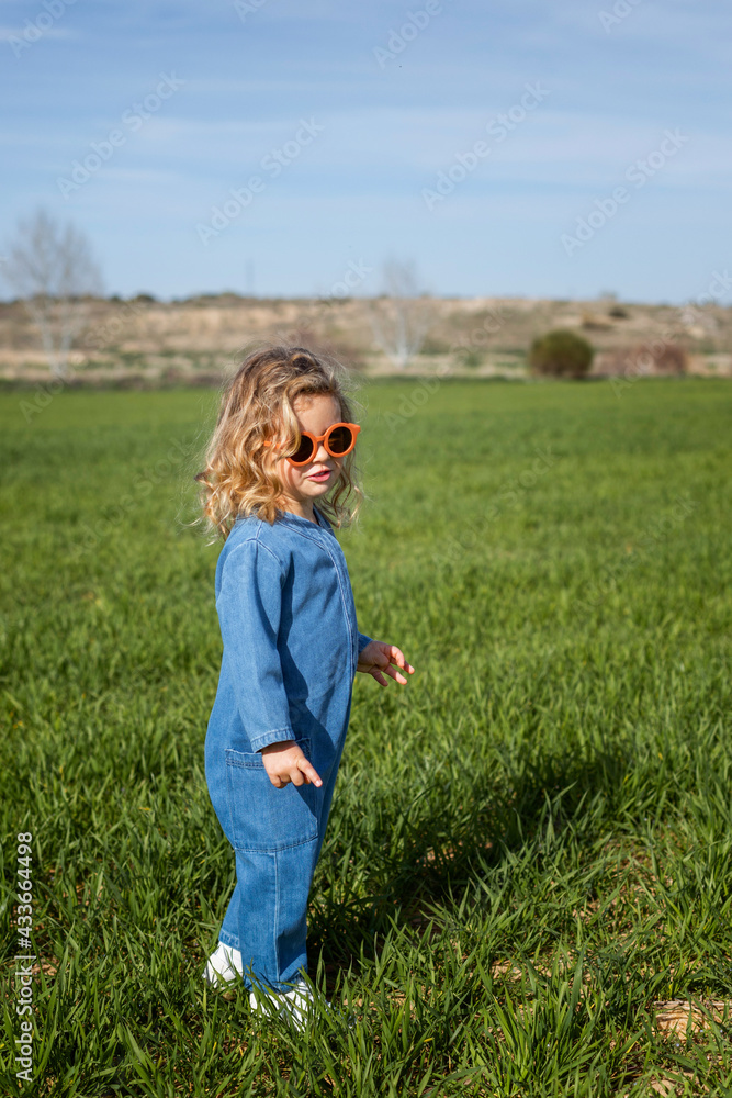 Side view of cute happy little girl in trendy clothes and sunglasses standing and relaxing on grassy lawn Side view of cute happy little girl in trendy clothes and sunglasses standing and relaxing on grassy lawn