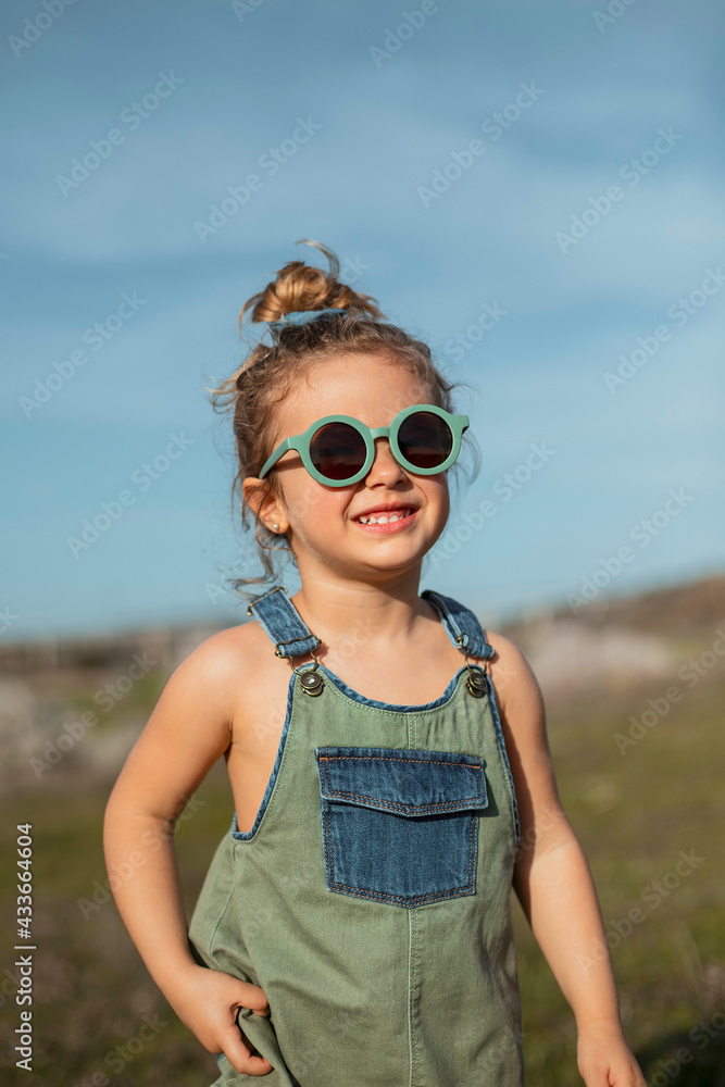 Content little girl in overalls and sunglasses standing in meadow and enjoying summer on sunny day in countryside Content little girl in overalls and sunglasses standing in meadow and enjoying summer on sunny day in countryside
