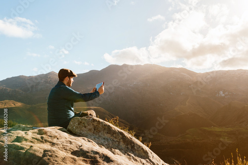 Side view of male explorer sitting on rocky hill and taking picture of mountain range while using tablet photo