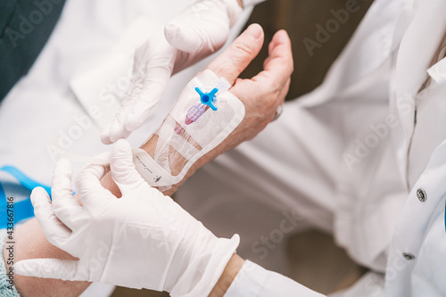 High angle of crop anonymous medic in disposable gloves putting intravenous catheter on arm of patient in hospital