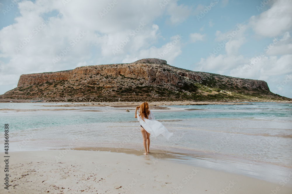 Full length slim female in black swimsuit standing on sandy Balos beach against rocky cliff and looking away on clear sunny weather Full length slim female in black swimsuit standing on sandy Balos beach against rocky cliff and looking away on clear sunny weather