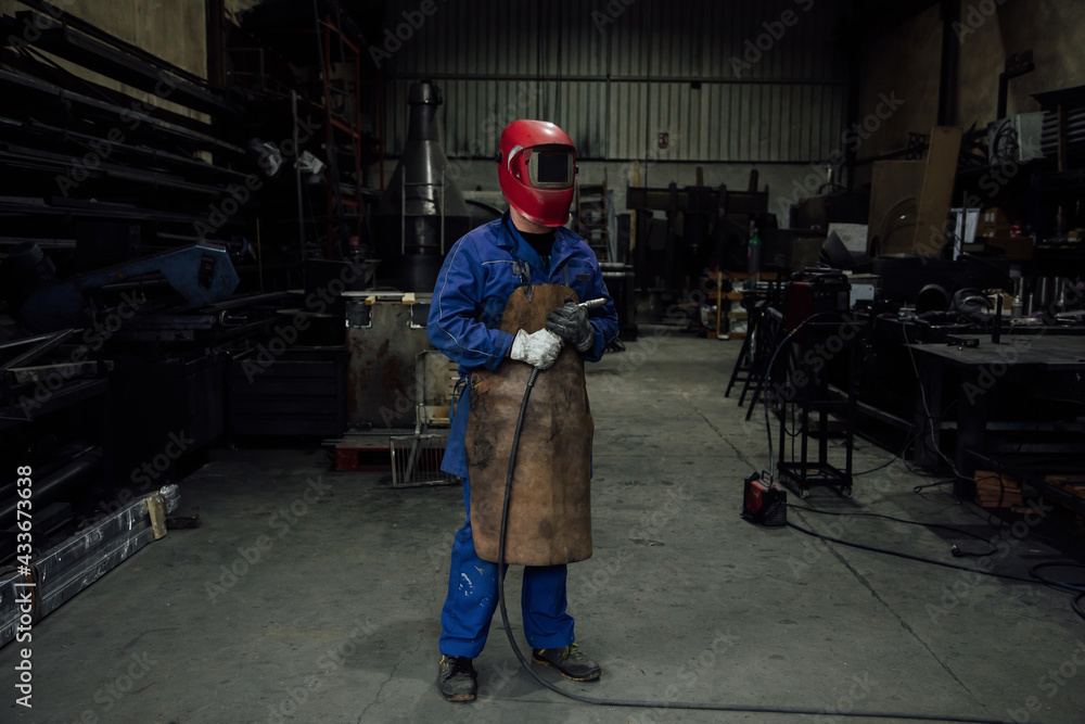 Anonymous mechanic in protective helmet and apron on blue overall standing with welding hose in light workshop near metal constructions Anonymous mechanic in protective helmet and apron on blue overall standing with welding hose in light workshop near metal constructions