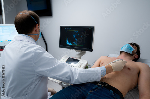 Male medic using modern ultrasound equipment and examining heart of male patient in mask lying on medical table during diagnostic in hospital photo