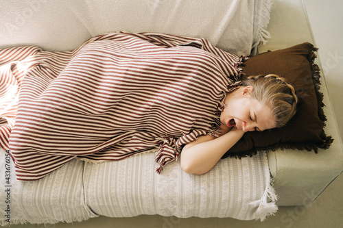 From above of young sleepy female yawning while lying under plaid on soft sofa at home
