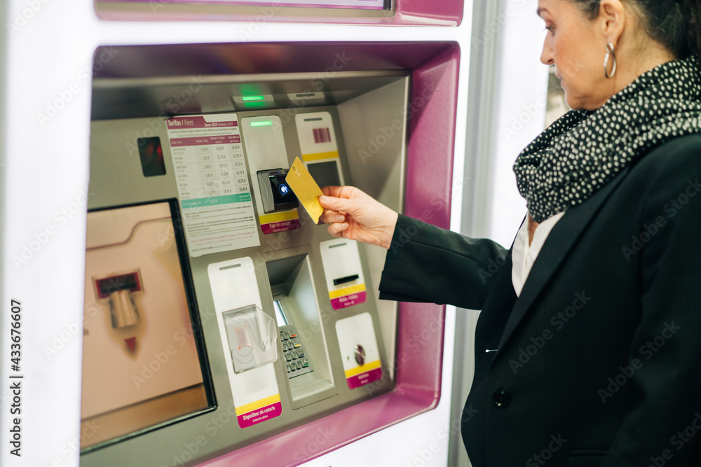 Side view of serious female standing near ATM machine with card while buying train ticket while standing in modern terminal Side view of serious female standing near ATM machine with card while buying train ticket while standing in modern terminal