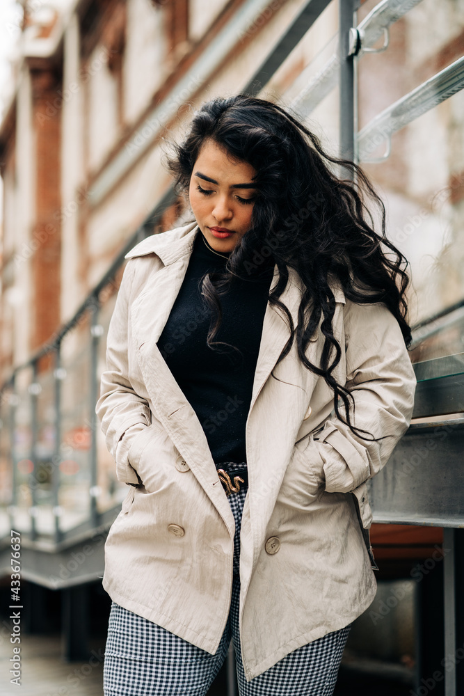 Low angle of plus size female wearing trendy coat standing with hands in pockets against city street and looking down Low angle of plus size female wearing trendy coat standing with hands in pockets against city street and looking down