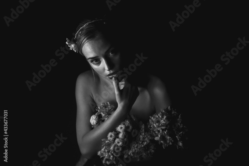 High angle black and white of charming young bare shouldered female with bunch of flowers and floral adornment on head touching lips and looking at camera in dark studio photo