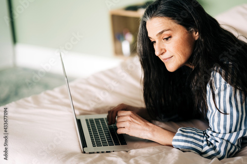 Side view of focused mature self employed Hispanic woman with long dark hair in casual clothes lying on bed and typing on laptop during online work at home