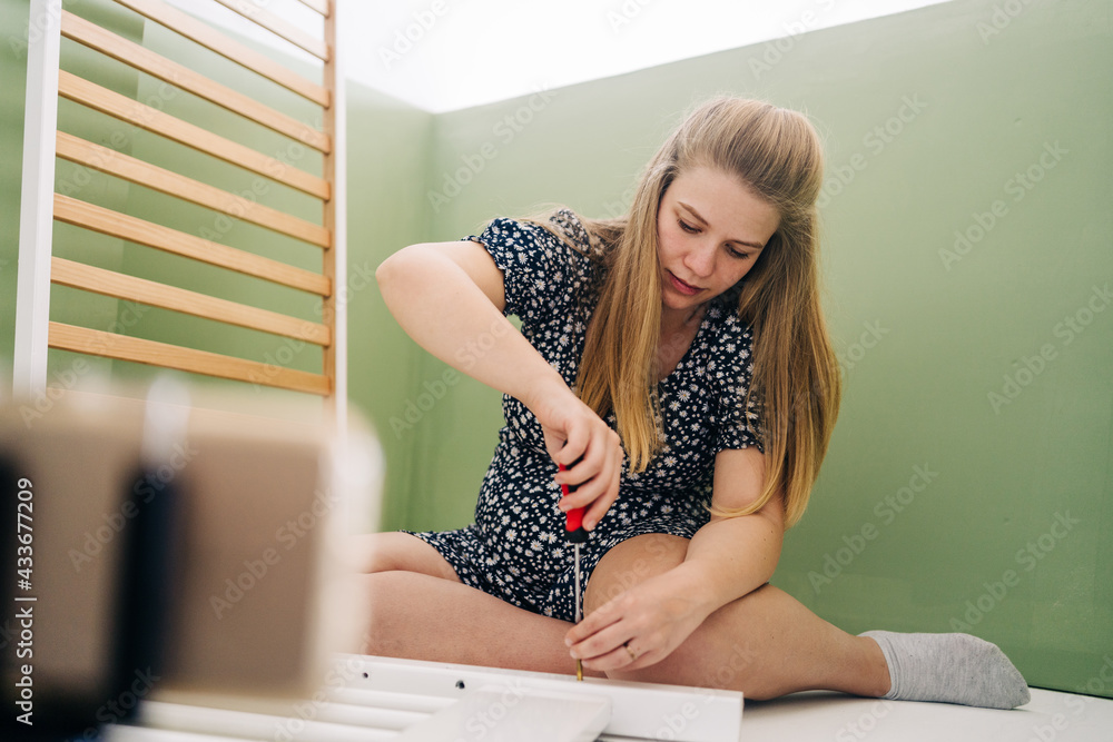 © Jake Jakab/ADDICTIVE STOCK - Young expectant female using screwdriver while assembling baby cot and sitting on floor at home