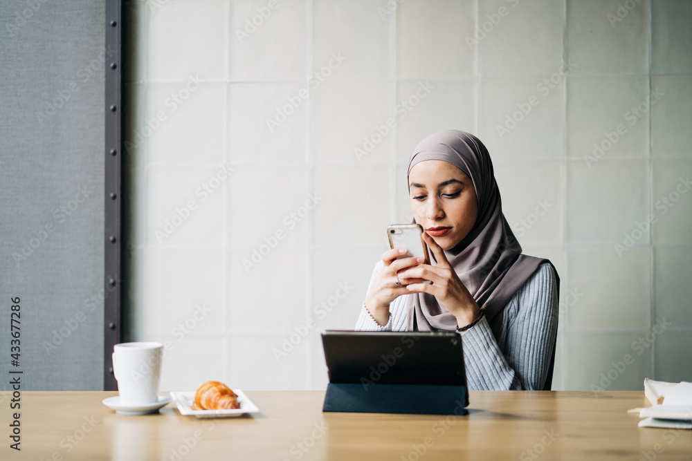 Side view of Muslim female freelancer wearing traditional hijab standing in cafe and browsing smartphone while working on project remotely Side view of Muslim female freelancer wearing traditional hijab standing in cafe and browsing smartphone while working on project remotely