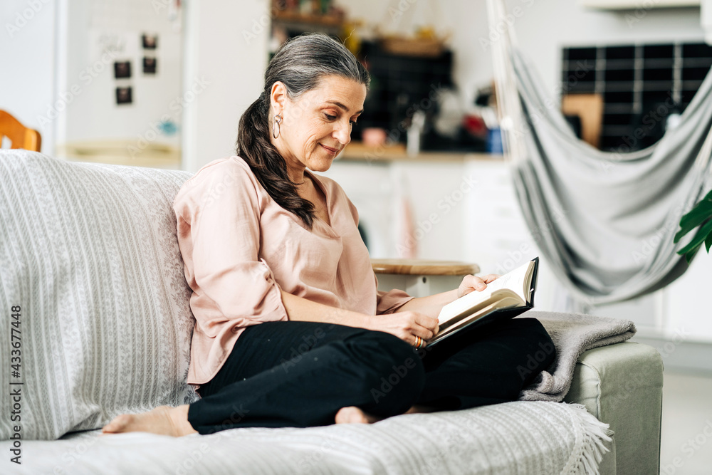 Full body of barefoot female reading book while sitting on comfortable sofa in living room with green plant at home Full body of barefoot female reading book while sitting on comfortable sofa in living room with green plant at home