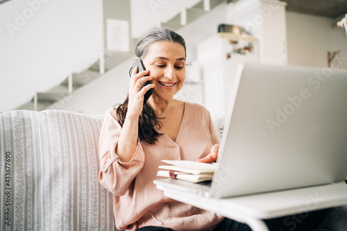 Concentrated middle aged female having phone call and taking notes in notepad while looking at screen of laptop in light room at home