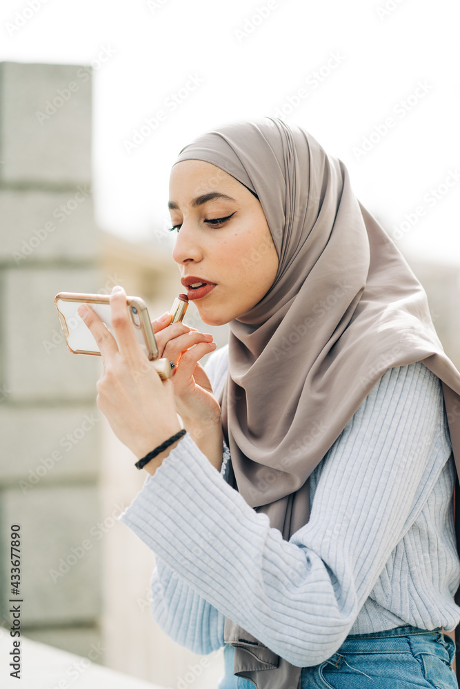 Side view of Muslim female in headscarf applying lipstick and looking at screen of smartphone while doing makeup in street Side view of Muslim female in headscarf applying lipstick and looking at screen of smartphone while doing makeup in street