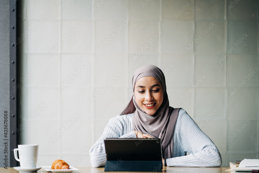 Content Muslim female in hijab and talking on video chat via tablet while sitting at table in cafe Content Muslim female in hijab and talking on video chat via tablet while sitting at table in cafe